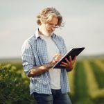 Agricultor idoso faz anotações em um caderno enquanto observa uma plantação.