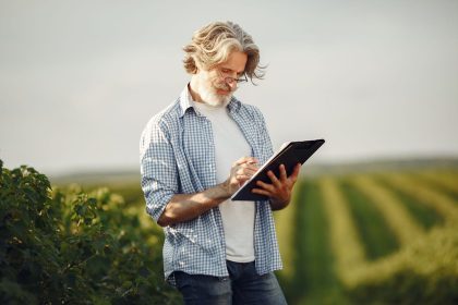 Agricultor idoso faz anotações em um caderno enquanto observa uma plantação.