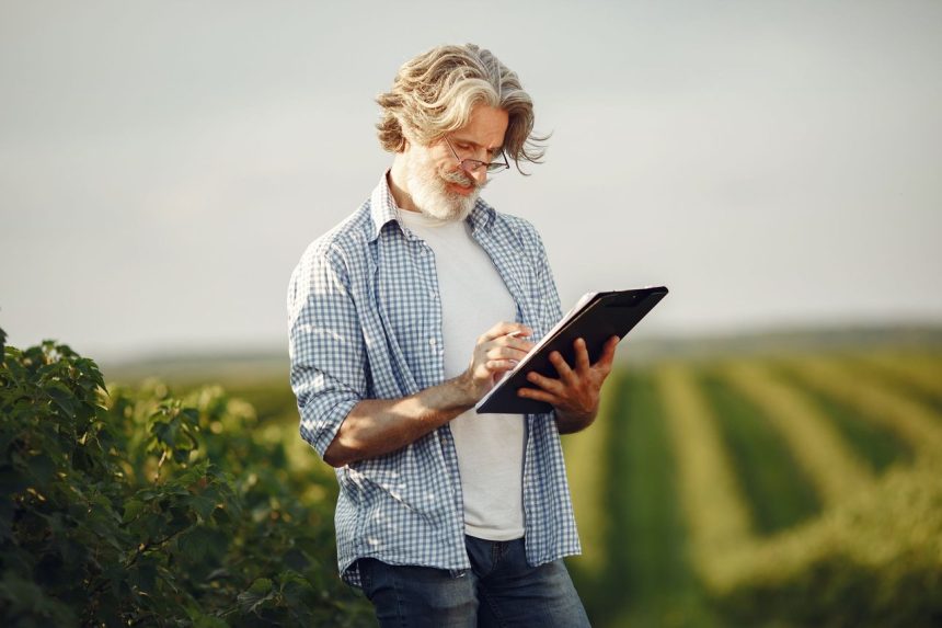 Agricultor idoso faz anotações em um caderno enquanto observa uma plantação.